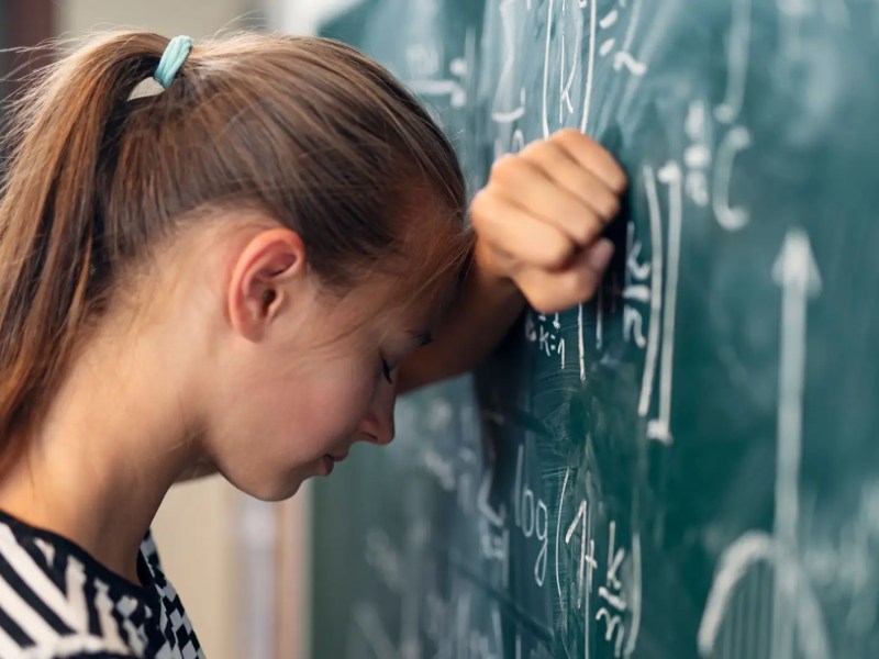 Mädchen lehnt mit Kopf an Tafel, auf der eine Matheaufgabe steht.