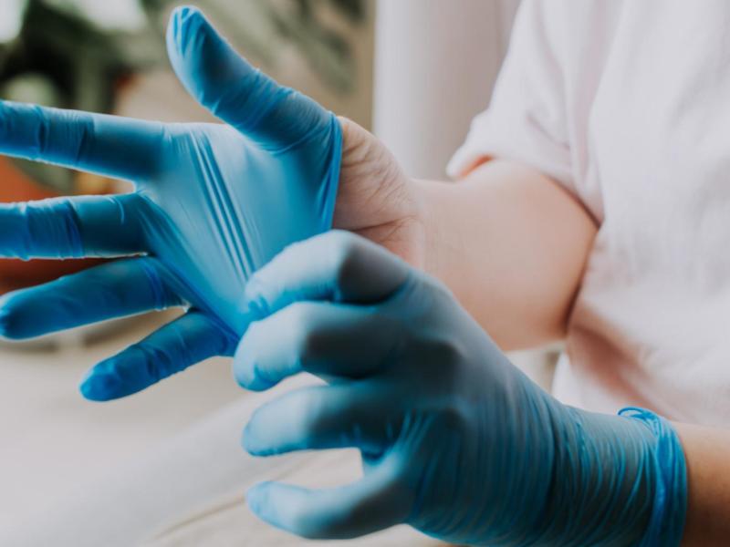 Eine Person mit hellem Shirt streift sich konzentriert blaue Einweghandschuhe &uuml;ber, im Hintergrund unscharf eine gr&uuml;ne Zimmerpflanze.