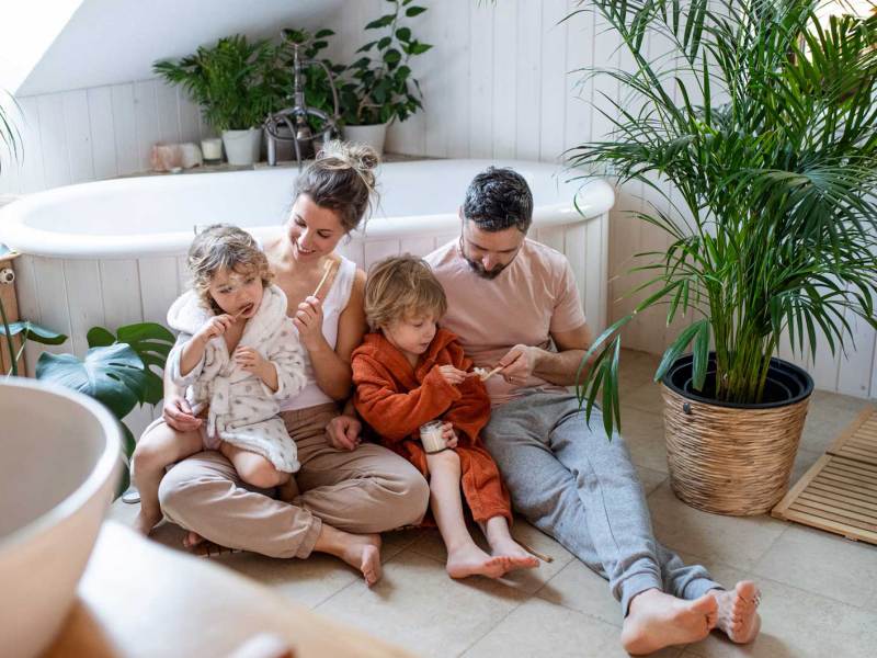 Familie sitzt auf dem Badezimmerboden und die Eltern helfen den Kindern beim Zähneputzen.