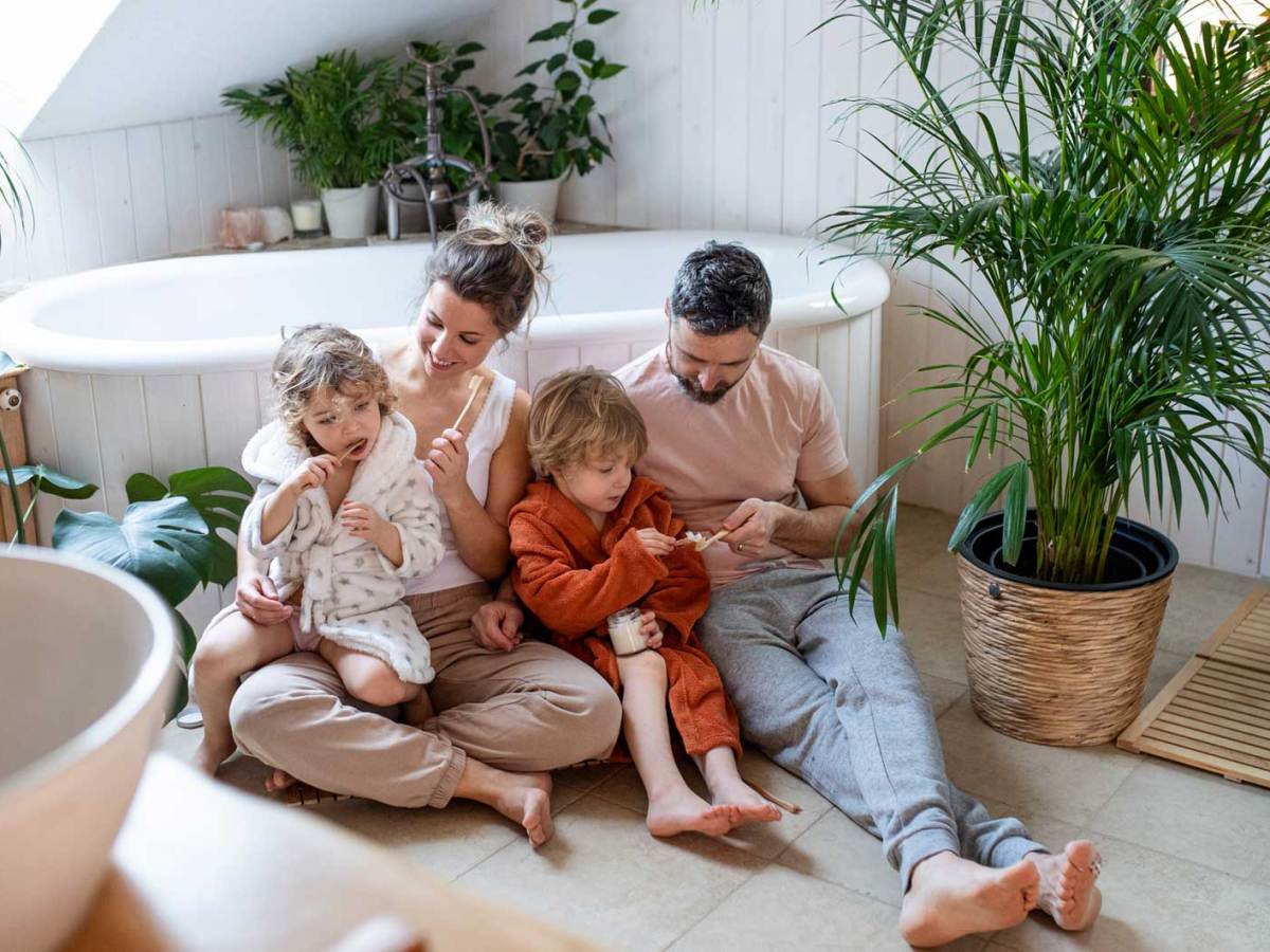 Familie sitzt auf dem Badezimmerboden und die Eltern helfen den Kindern beim Zähneputzen.