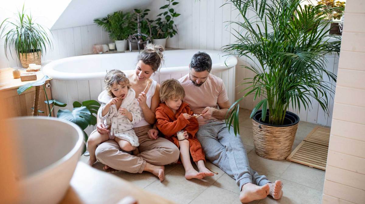 Familie sitzt auf dem Badezimmerboden und die Eltern helfen den Kindern beim Zähneputzen.