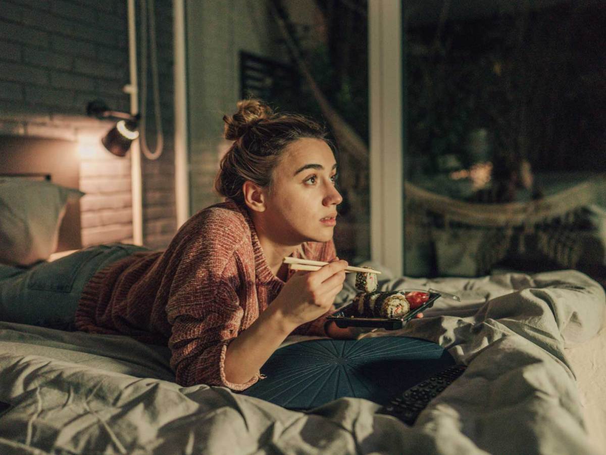 Frau liegt auf dem Bett, isst Sushi und schaut etwas Spannendes.