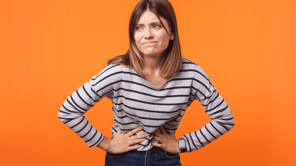 Portr&auml;t einer ungl&uuml;cklichen, kranken Frau mit braunen Haaren in einem lang&auml;rmeligen gestreiften Hemd, die steht und sich mit den H&auml;nden den Bauch h&auml;lt, Bauchkr&auml;mpfe oder Regelschmerzen hat.