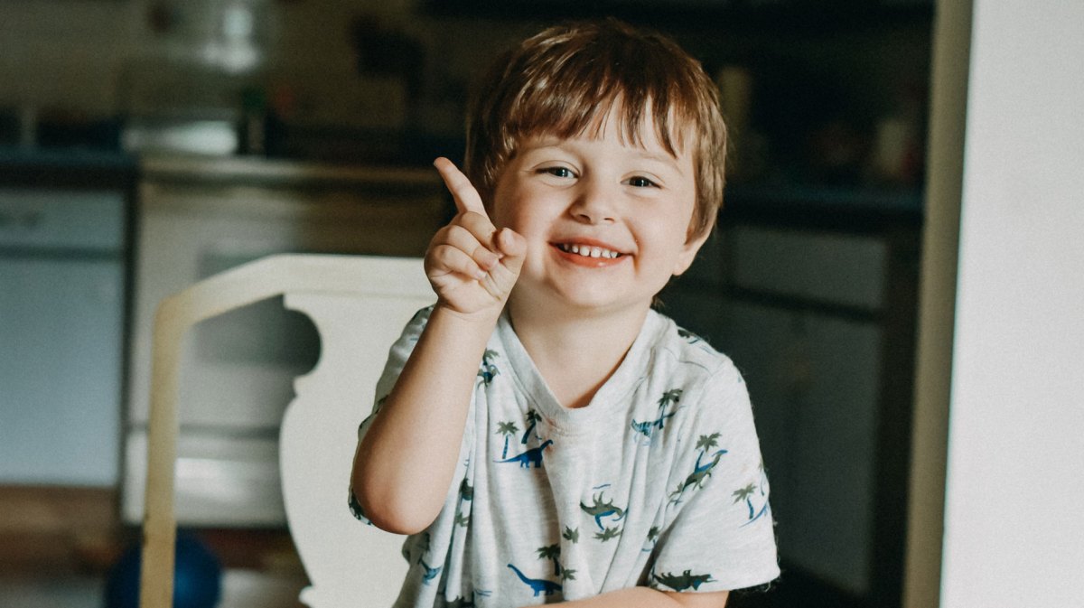 Kind am Küchentisch hebt den Finger und möchte etwas sagen.