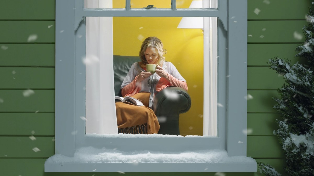 Junge blonde Frau sitzt lesend mit Tee und Decke auf einem Ledersessel hinter vereistem Fenster, draußen fällt Schnee.