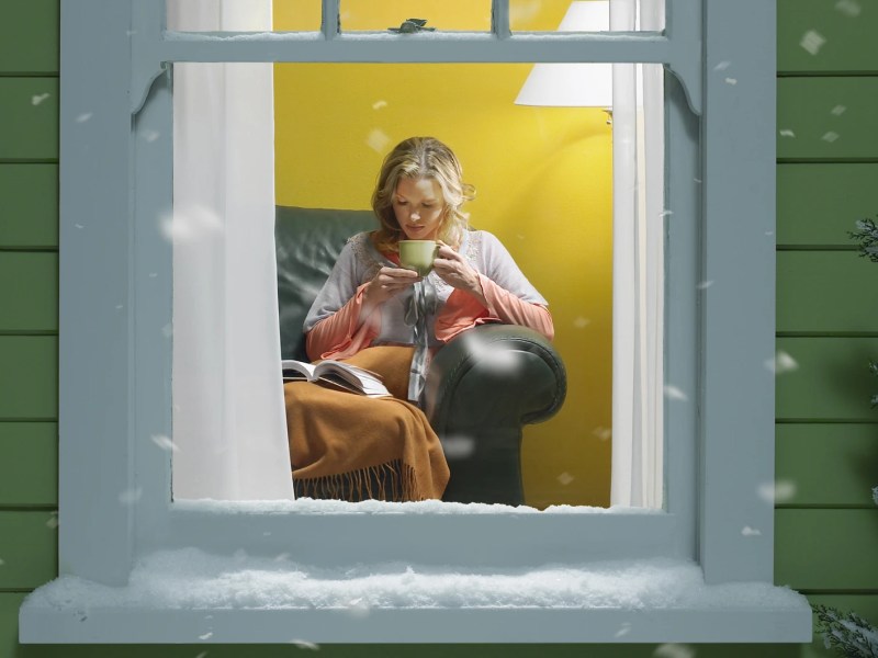 Junge blonde Frau sitzt lesend mit Tee und Decke auf einem Ledersessel hinter vereistem Fenster, draußen fällt Schnee.