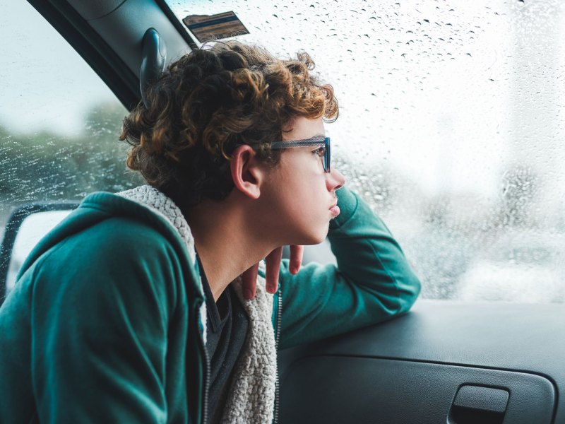 Junge im Teenie-Alter sitzt an einem Regentag im Auto und schaut sehns&uuml;chtig aus dem Fenster.