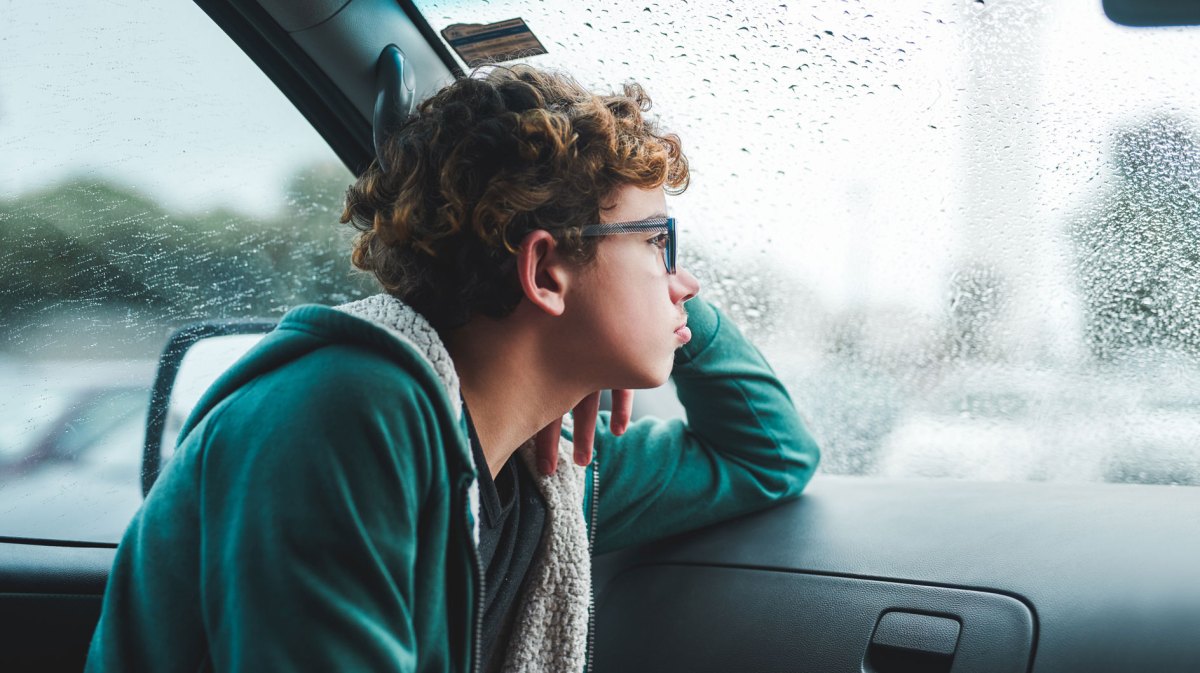 Junge im Teenie-Alter sitzt an einem Regentag im Auto und schaut sehnsüchtig aus dem Fenster.