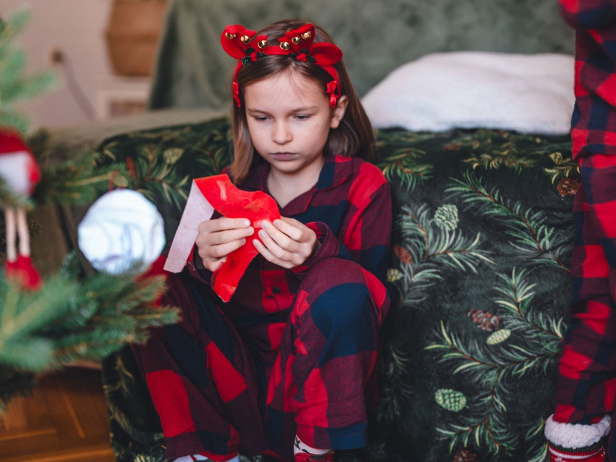 M&auml;dchen sitzt am Weihnachtsmorgen entt&auml;uscht neben dem Weihnachtsbaum und packt ein Geschenk aus.