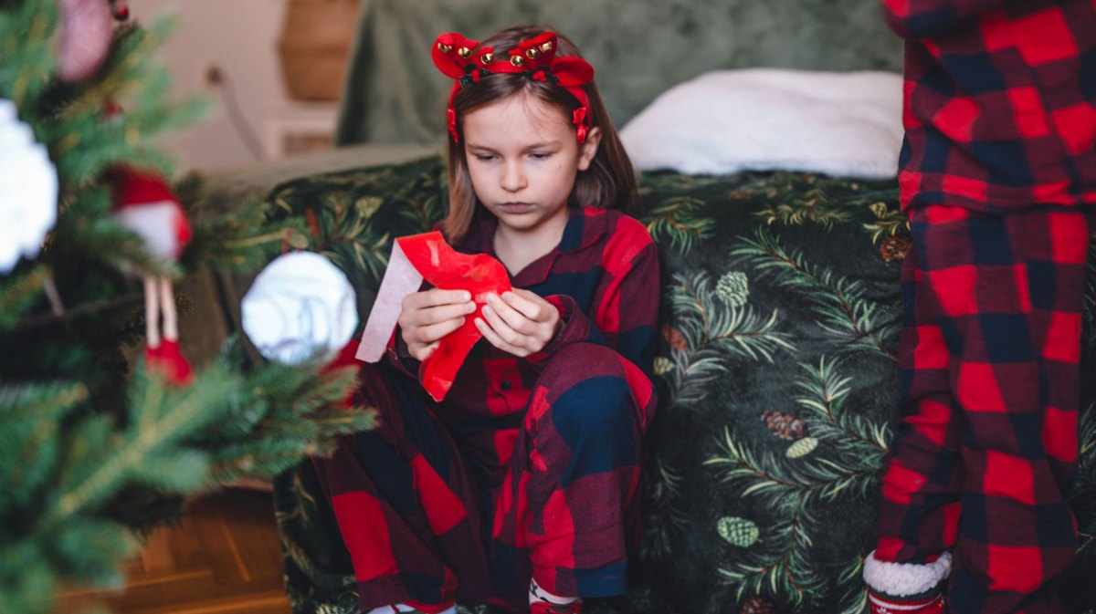 Mädchen sitzt am Weihnachtsmorgen enttäuscht neben dem Weihnachtsbaum und packt ein Geschenk aus.