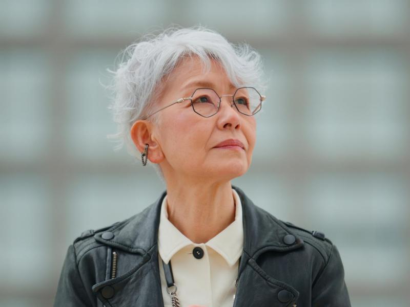 Junggebliebene &auml;ltere Frau mit wei&szlig;en Haaren und Lederjacke und Brille und guckt zur Seite.