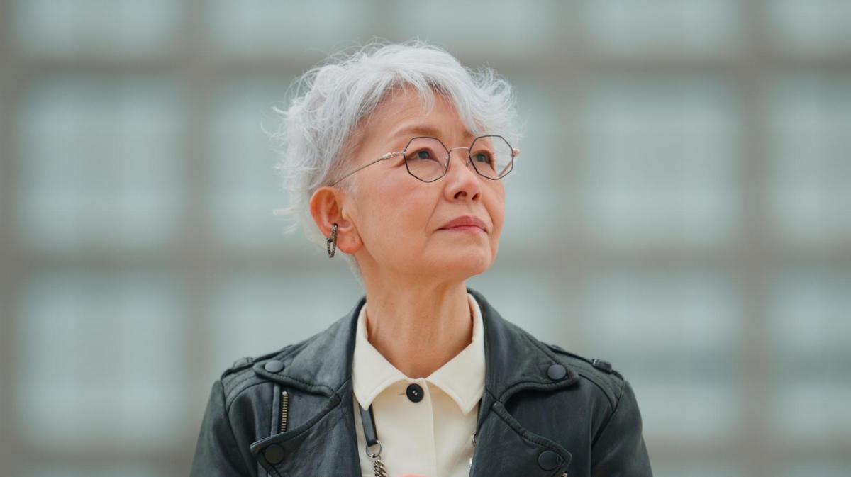 Junggebliebene ältere Frau mit weißen Haaren und Lederjacke und Brille und guckt zur Seite.