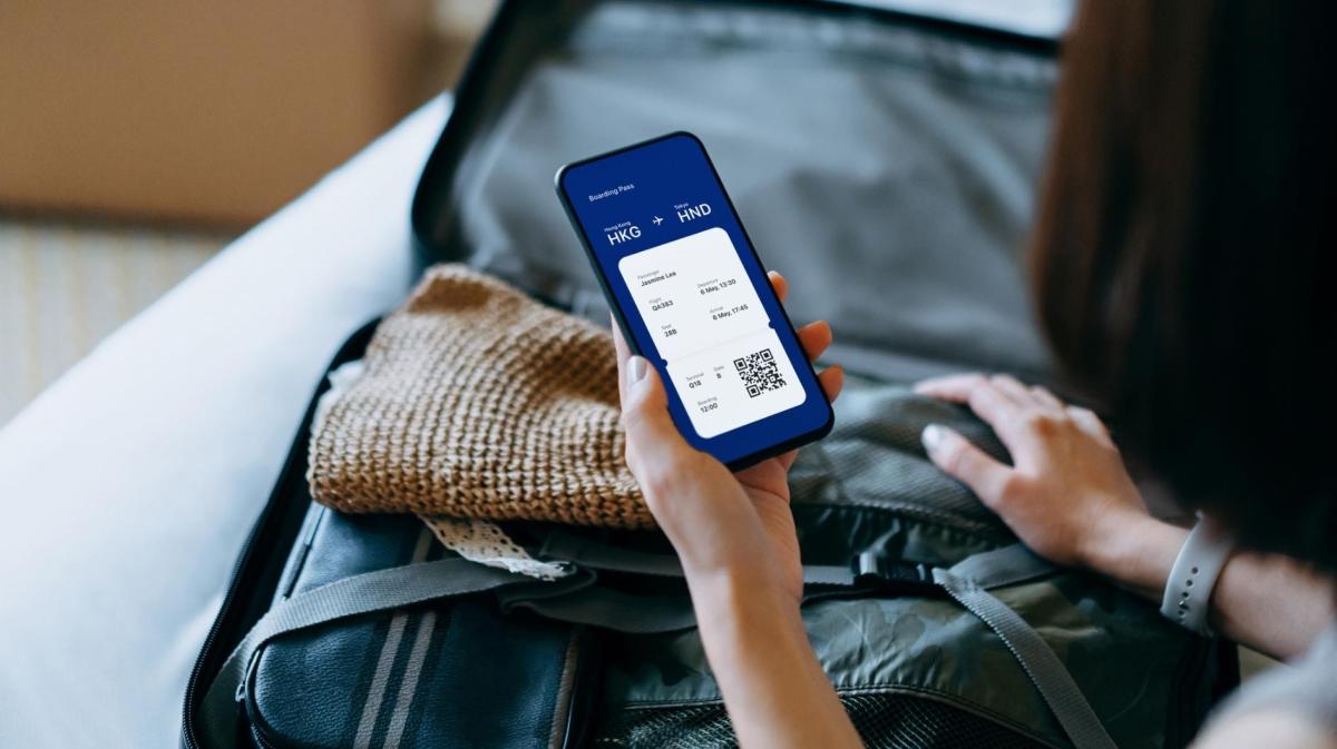 Frau sitzt mit einem gepackten Koffer und einem Handy auf dem Bett.