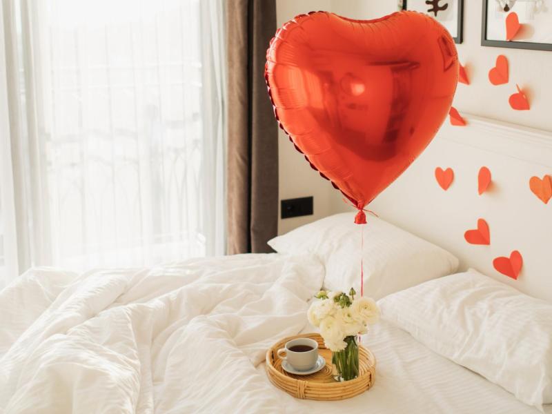 Roter Herzballon auf einem Bett mit wei&szlig;er Bettw&auml;sche, auf dem ein kleines Tablett mit einem Kaffee steht.