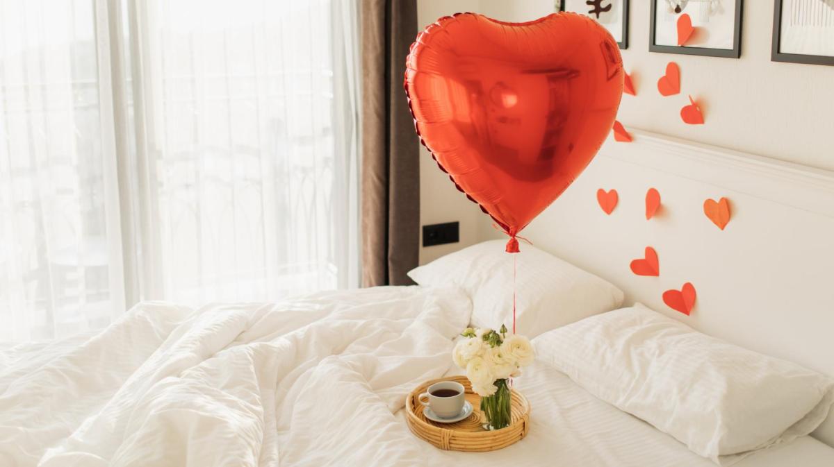Roter Herzballon auf einem Bett mit weißer Bettwäsche, auf dem ein kleines Tablett mit einem Kaffee steht.