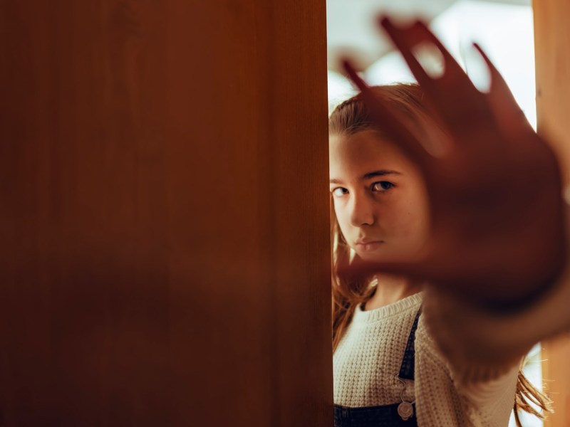 M&auml;dchen im Teenageralter steht an einer halb ge&ouml;ffneten T&uuml;r und zeigt mit der Hand Stopp an.