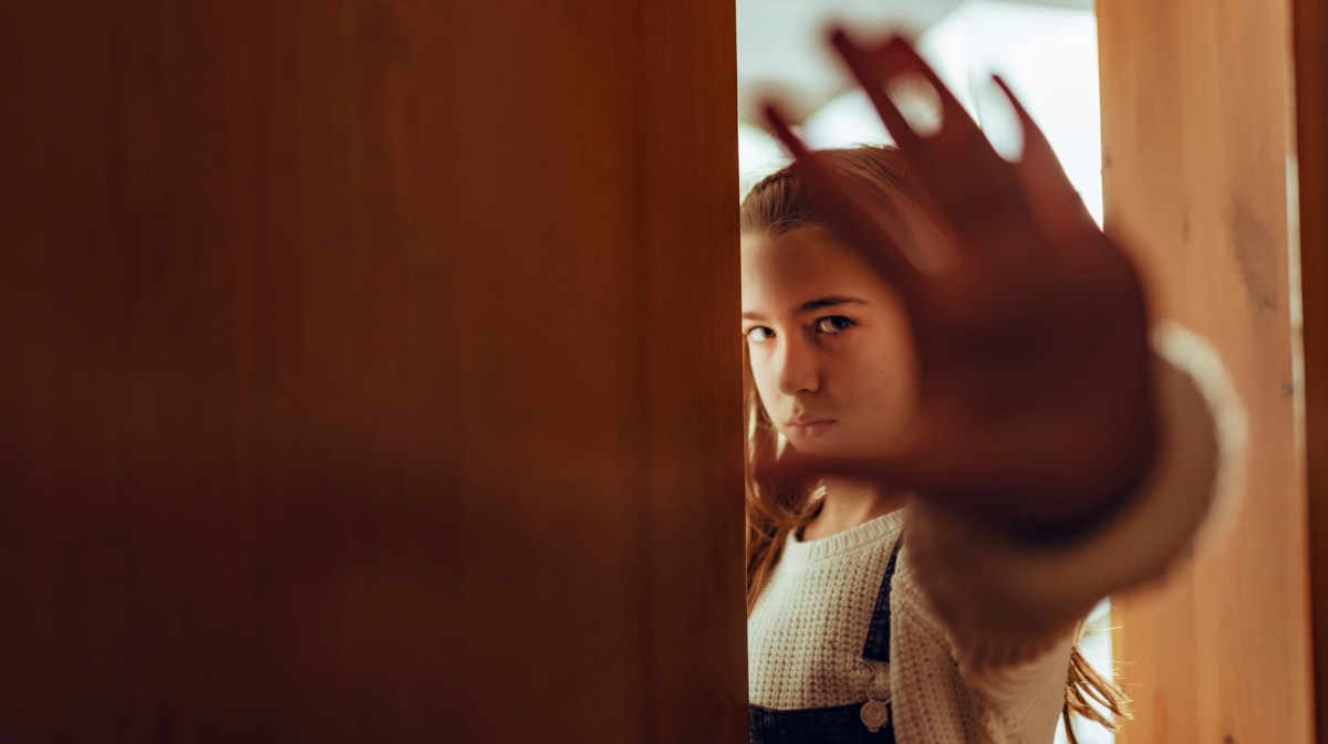 Mädchen im Teenageralter steht an einer halb geöffneten Tür und zeigt mit der Hand Stopp an.