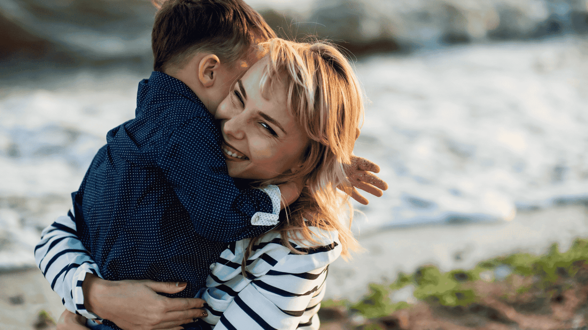 Frau mit blonden Haaren umarmt lächelnd einen kleinen Jungen.