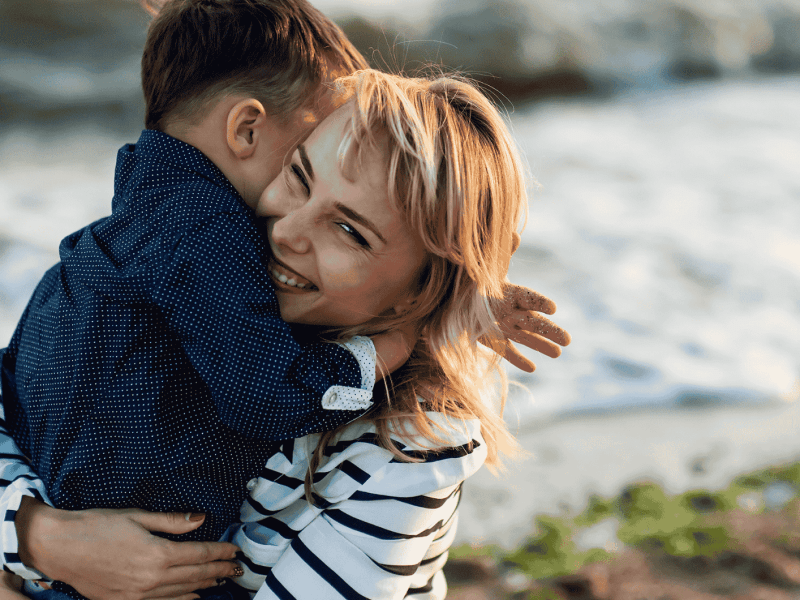 Frau mit blonden Haaren umarmt lächelnd einen kleinen Jungen.