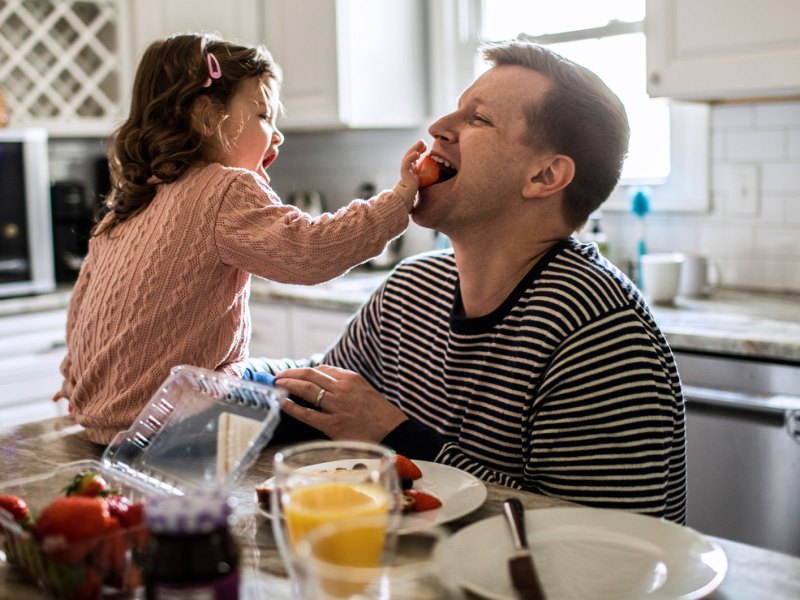 Tochter f&uuml;ttert ihren Papa am Fr&uuml;hst&uuml;ckstisch mit Erdbeeren.