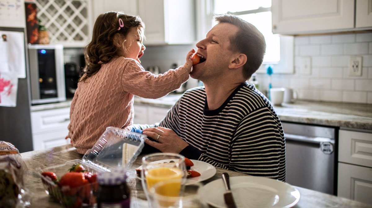 Tochter füttert ihren Papa am Frühstückstisch mit Erdbeeren.
