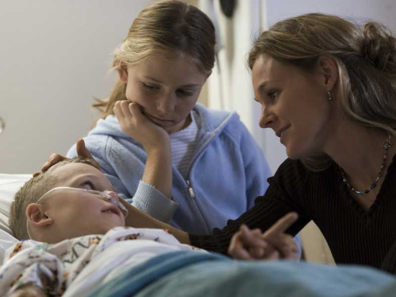 Mutter und &auml;lteres Geschwisterkind sitzen am Krankenbett eines Kleinkindes