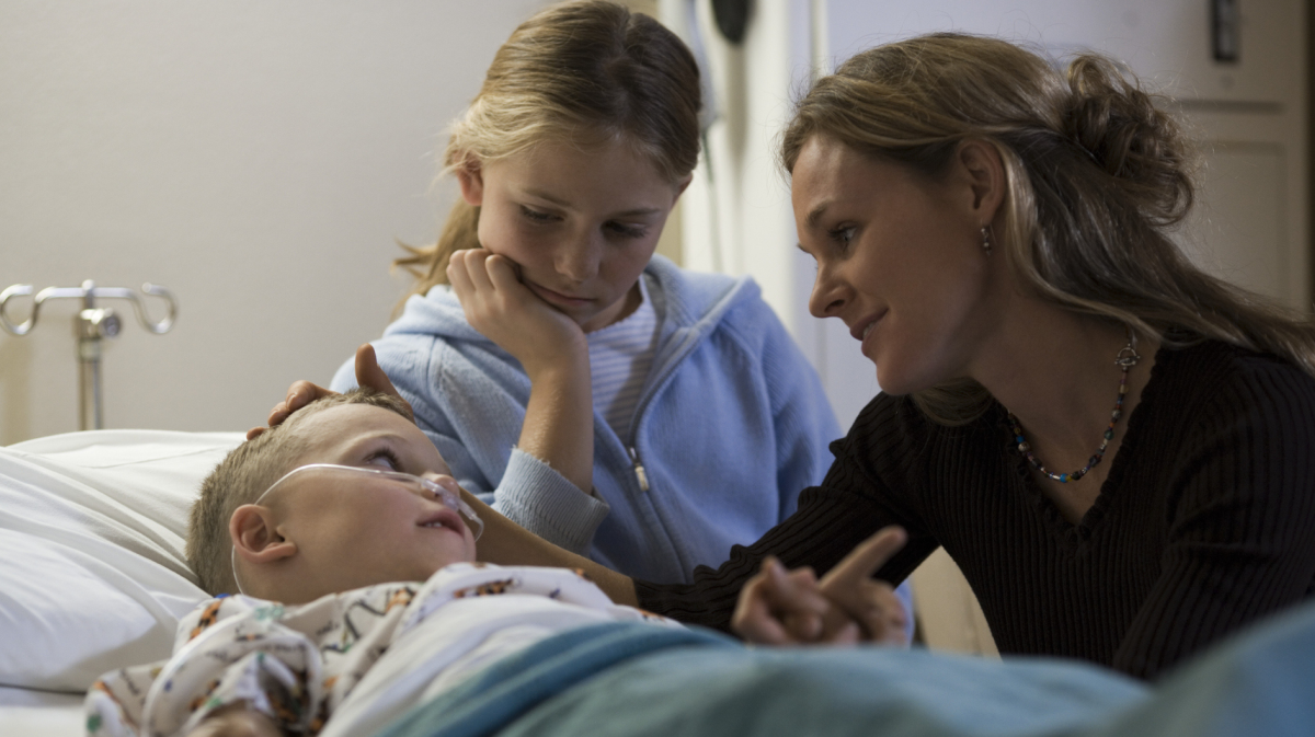 Mutter und älteres Geschwisterkind sitzen am Krankenbett eines Kleinkindes