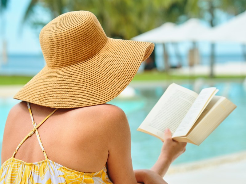 Frau mit Sonnenhut am Pool, die ein Buch liest