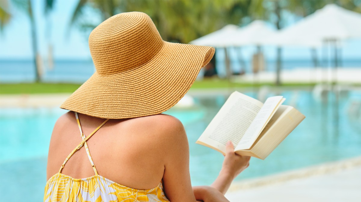 Frau mit Sonnenhut am Pool, die ein Buch liest