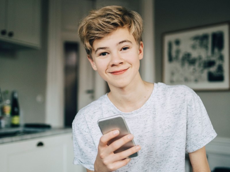 Teenagerjunge, ca 14 Jahre, mit Handy in der Hand, lacht in die Kamera.
