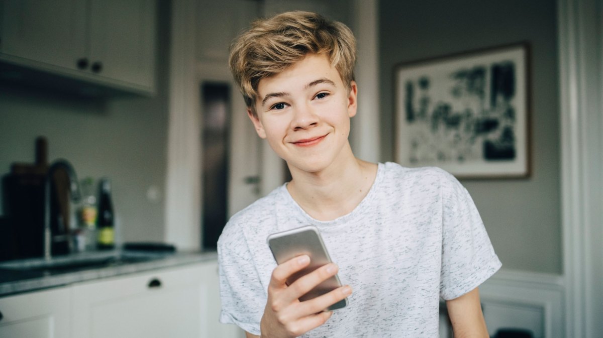 Teenagerjunge, ca 14 Jahre, mit Handy in der Hand, lacht in die Kamera.