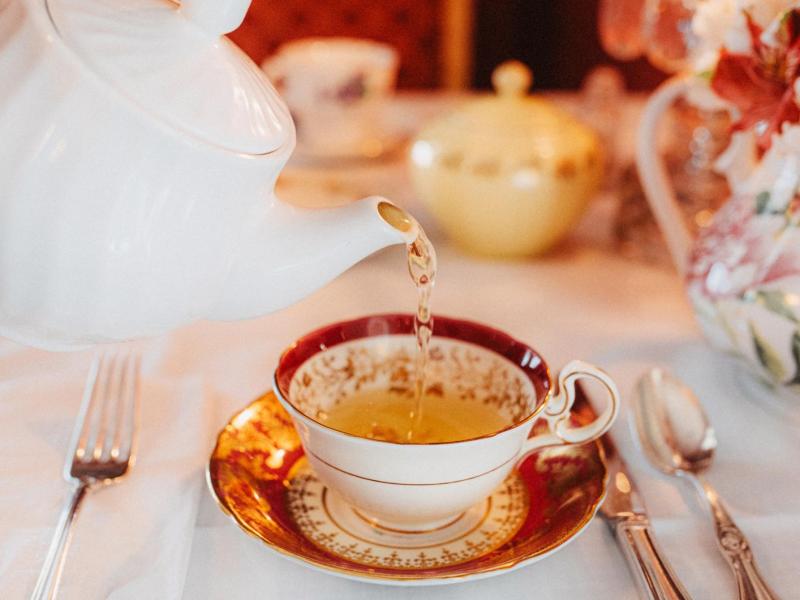 Person gie&szlig;t auf Tee aus einer Porzellan-Teekanne in eine Porzellanteetasse auf einem edel gedeckten Tisch.