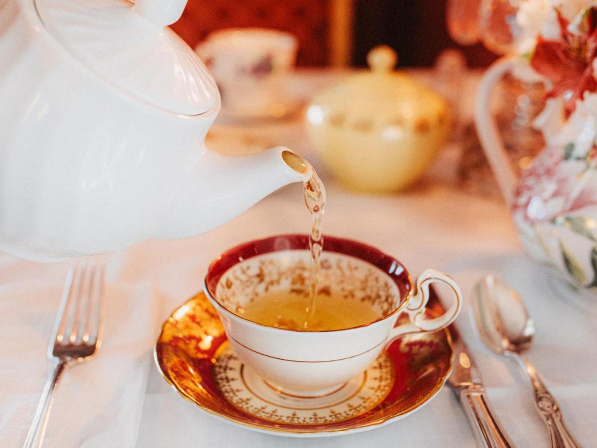 Person gie&szlig;t auf Tee aus einer Porzellan-Teekanne in eine Porzellanteetasse auf einem edel gedeckten Tisch.