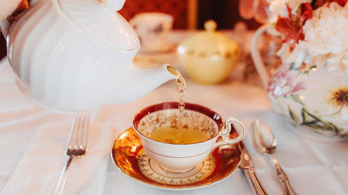 Person gießt auf Tee aus einer Porzellan-Teekanne in eine Porzellanteetasse auf einem edel gedeckten Tisch.