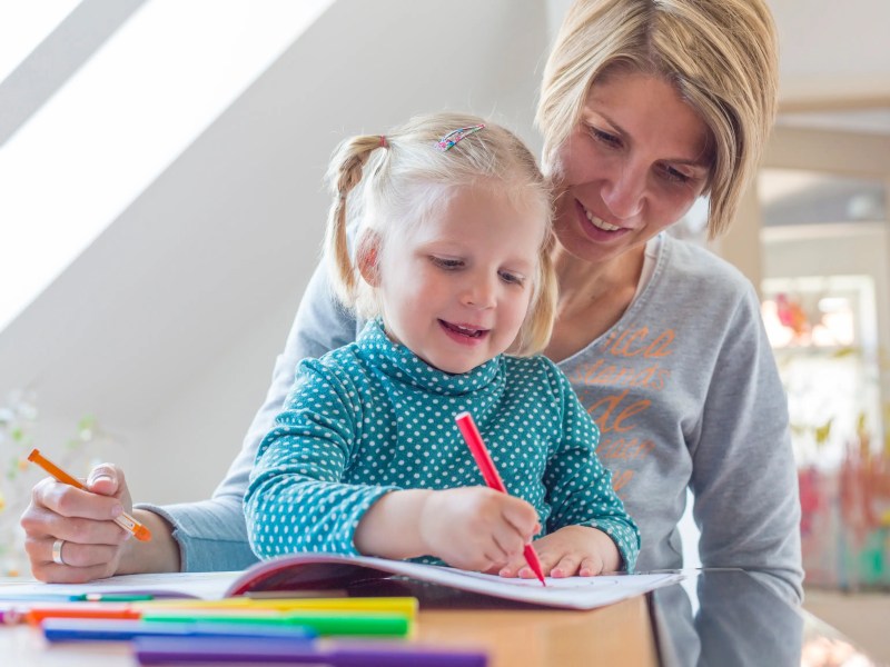 Junge Mutter sitzt mit ihrer Tochter auf dem Scho&szlig; am Tisch und malt.