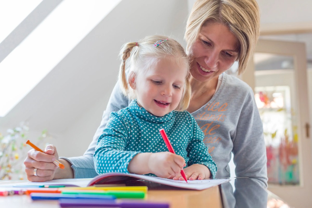 Junge Mutter sitzt mit ihrer Tochter auf dem Schoß am Tisch und malt.