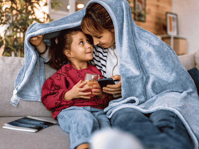 Mutter und Tochter kuscheln auf Sofa unter Kuscheldecke.