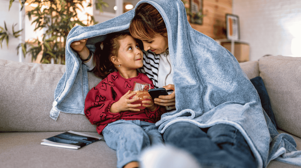 Mutter und Tochter kuscheln auf Sofa unter Kuscheldecke.