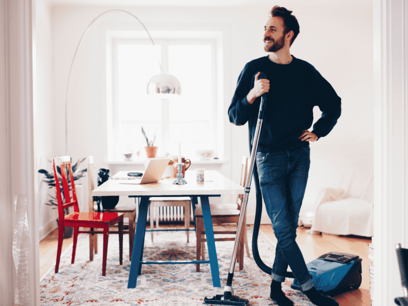Mann steht im Wohnzimmer, Staubsauger in der Hand.