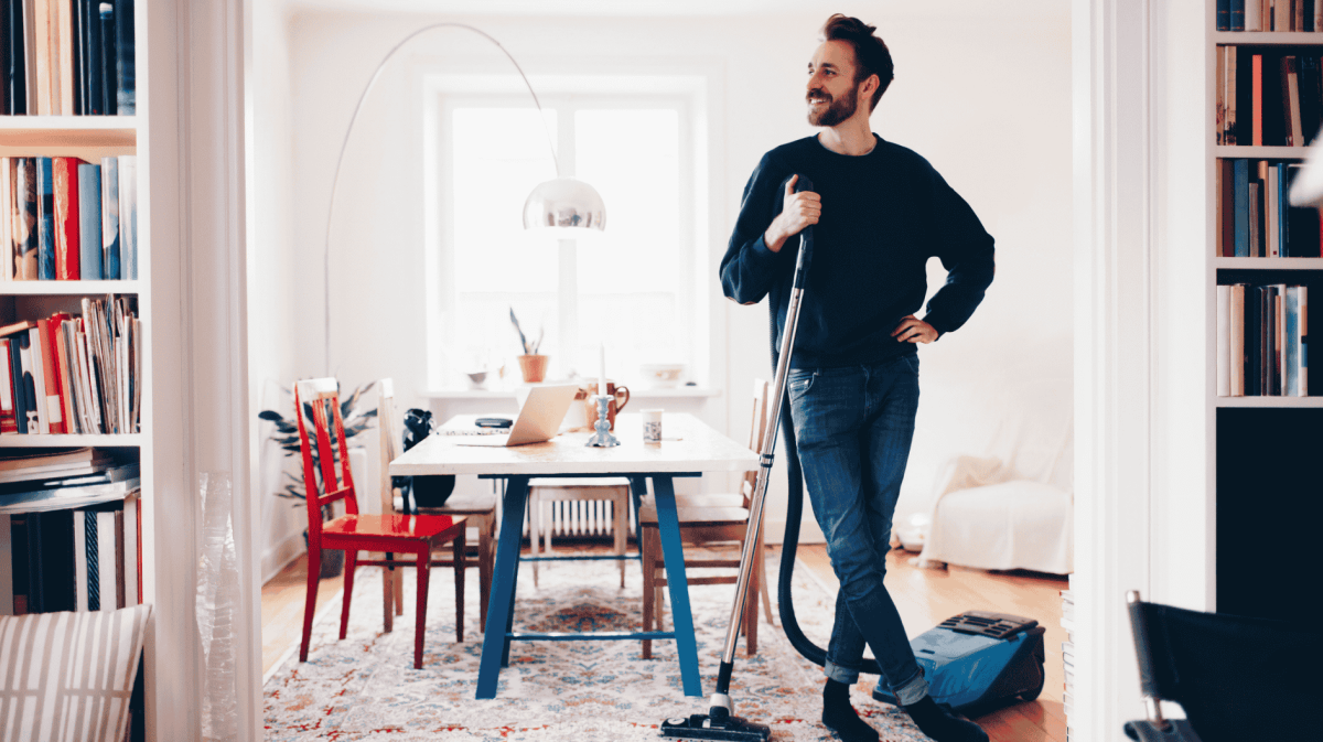 Mann steht im Wohnzimmer, Staubsauger in der Hand.