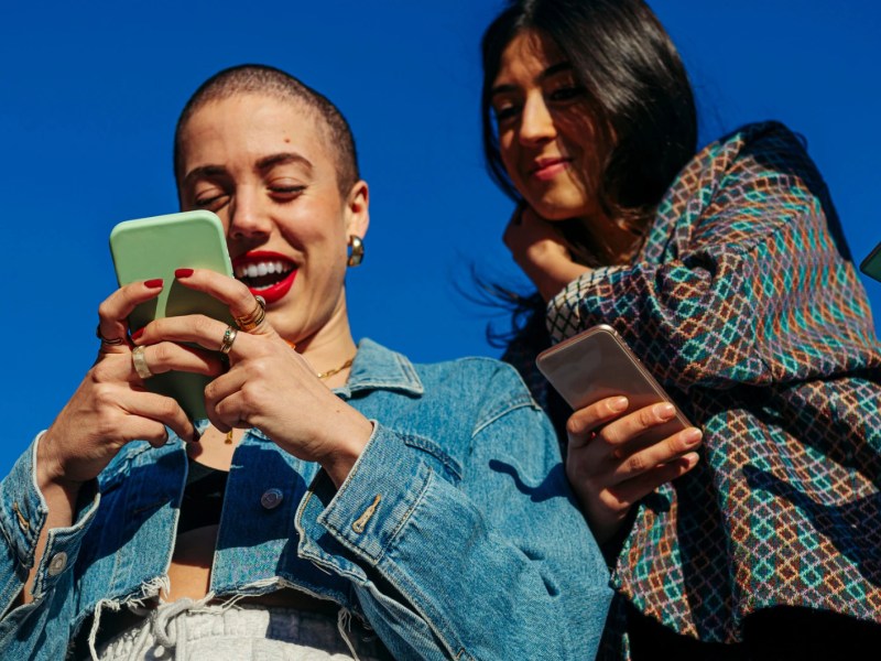 Freundinnen schauen auf ein Smartphone vor einem blauen Himmel