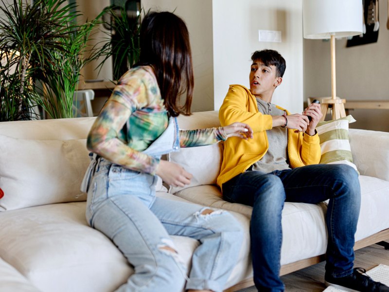 Bruder und Schwester sitzen nebeneinander auf dem Sofa und streiten um die Fernbedienung.