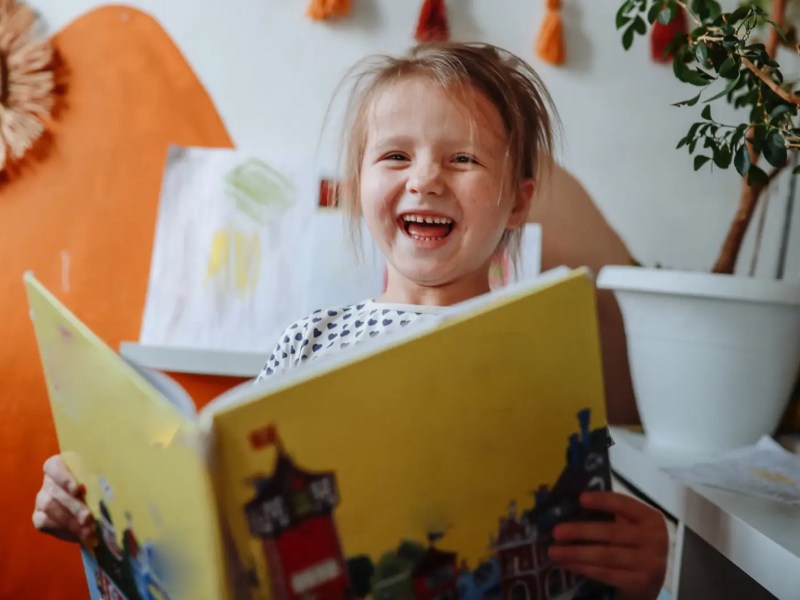 Mädchen, ca 4 Jahre, sitzt mit einem Buch in einem Kinderzimmer und lacht herzlich.