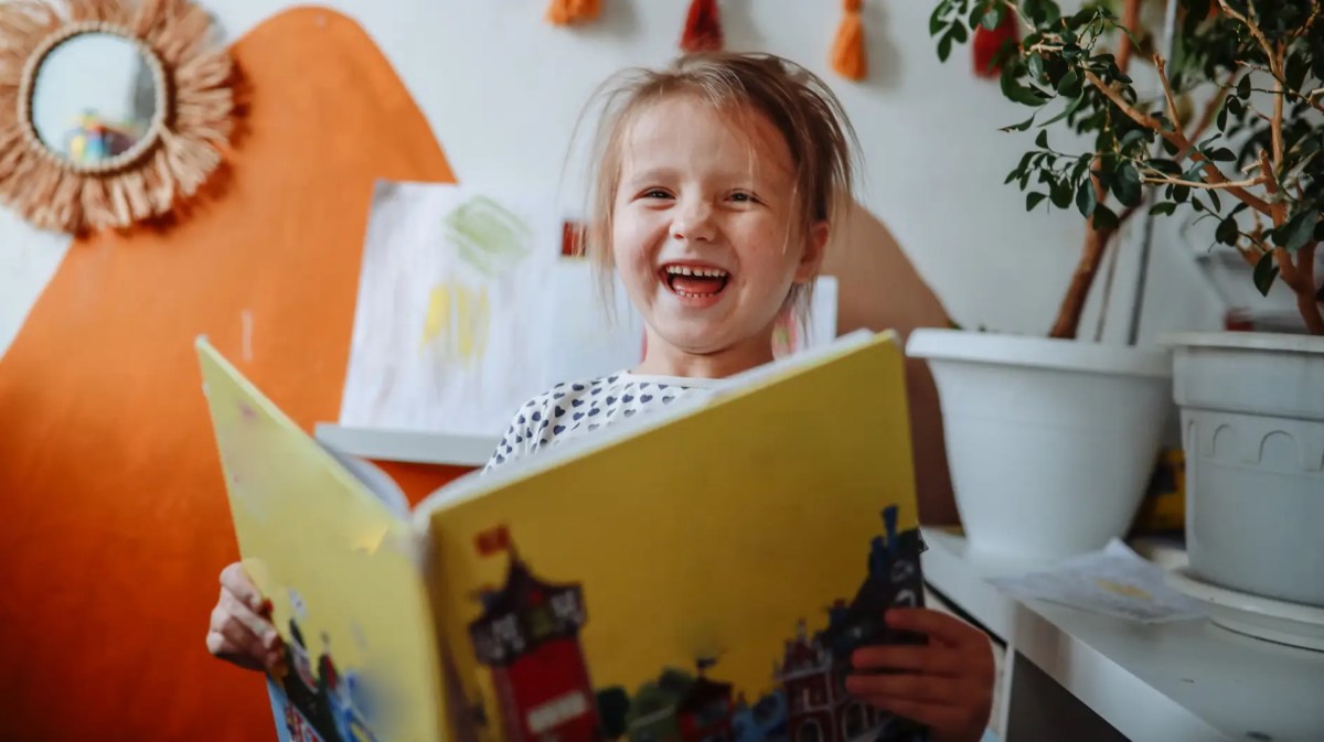 Mädchen, ca 4 Jahre, sitzt mit einem Buch in einem Kinderzimmer und lacht herzlich.