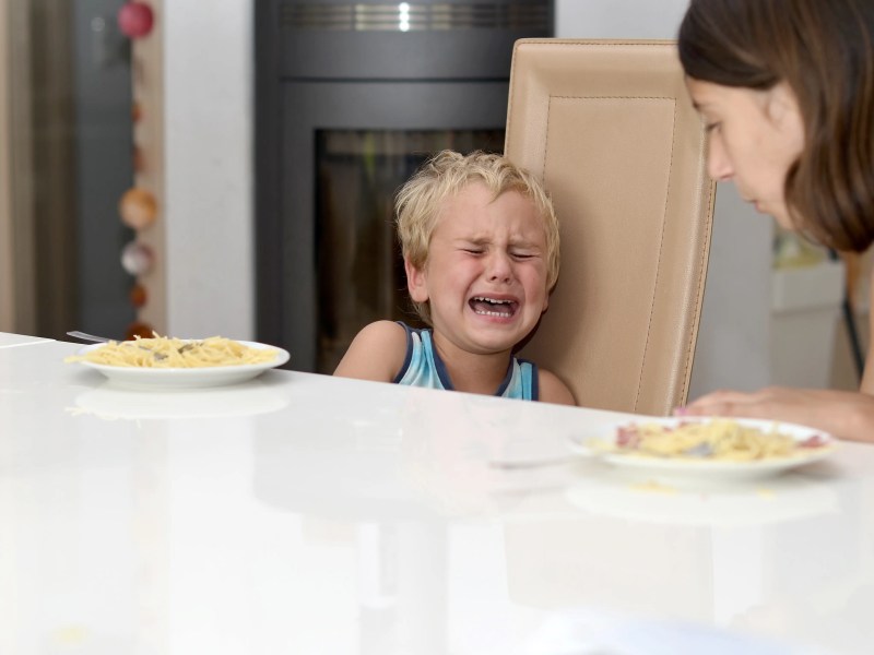 Junge sitzt schreiend am Esstisch mit seiner Mutter, dieihn versucht zu trösten.