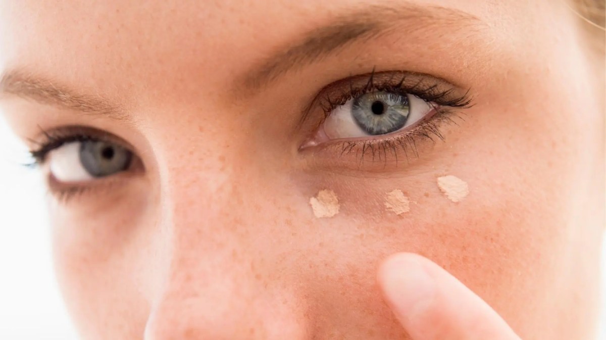 Frau mit hellen blauen Augen tupft sich Concealer unter das Auge.