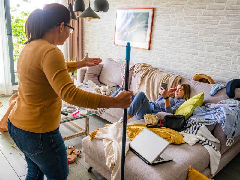 Mutter steht schimpfend vor ihrer Tochter, die im Chaos auf der Couch sitzt und am Handy ist.