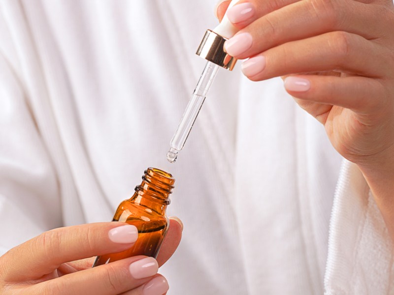 Beautiful Woman in white bathrobe holds glass bottle with pipette of Beauty Product Facial Serum or Essential Oil. Concept of home face and body self care and healthy lifestyle. Trendy beige pink nude manicure. Close-up. Copy space. Front view.
