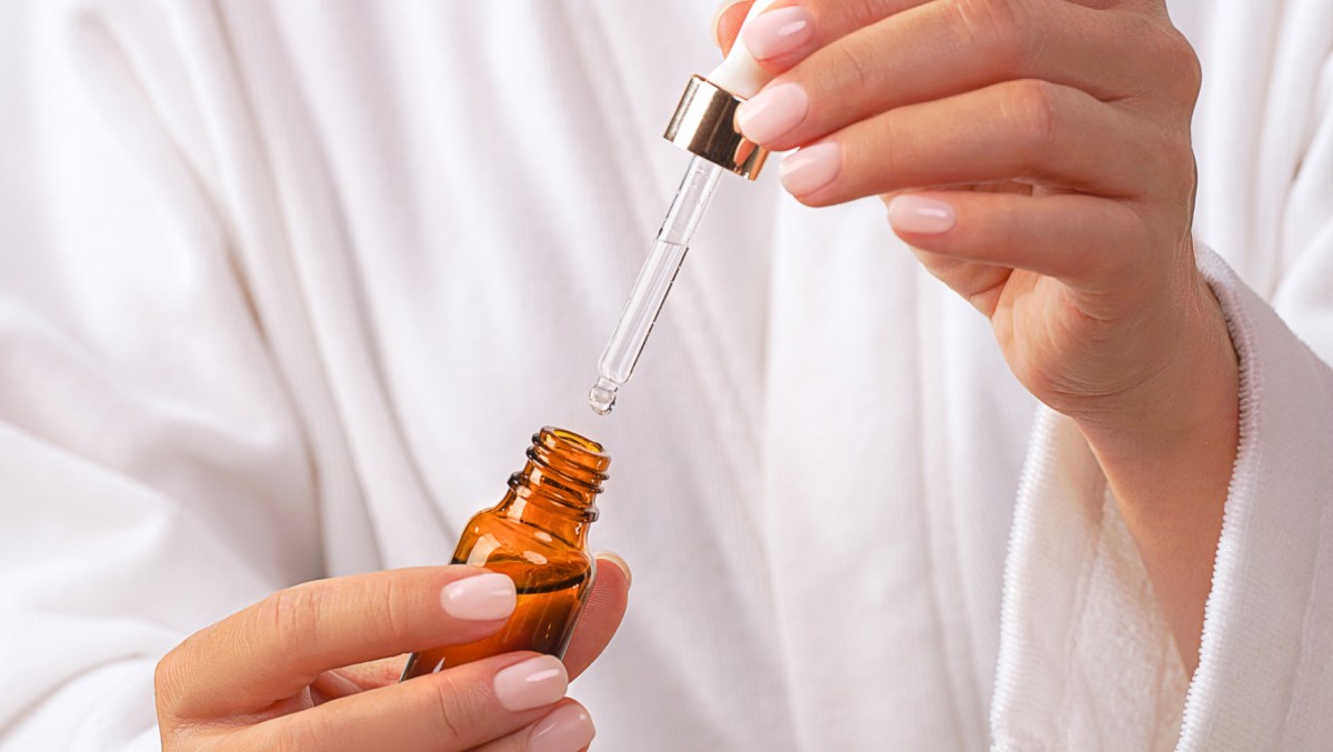 Beautiful Woman in white bathrobe holds glass bottle with pipette of Beauty Product Facial Serum or Essential Oil. Concept of home face and body self care and healthy lifestyle. Trendy beige pink nude manicure. Close-up. Copy space. Front view.