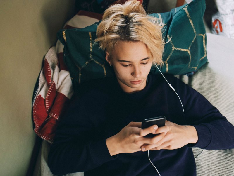 Teenager liegt auf dem Bett, das Handy in der Hand, Kopfh&ouml;rer im Ohr.